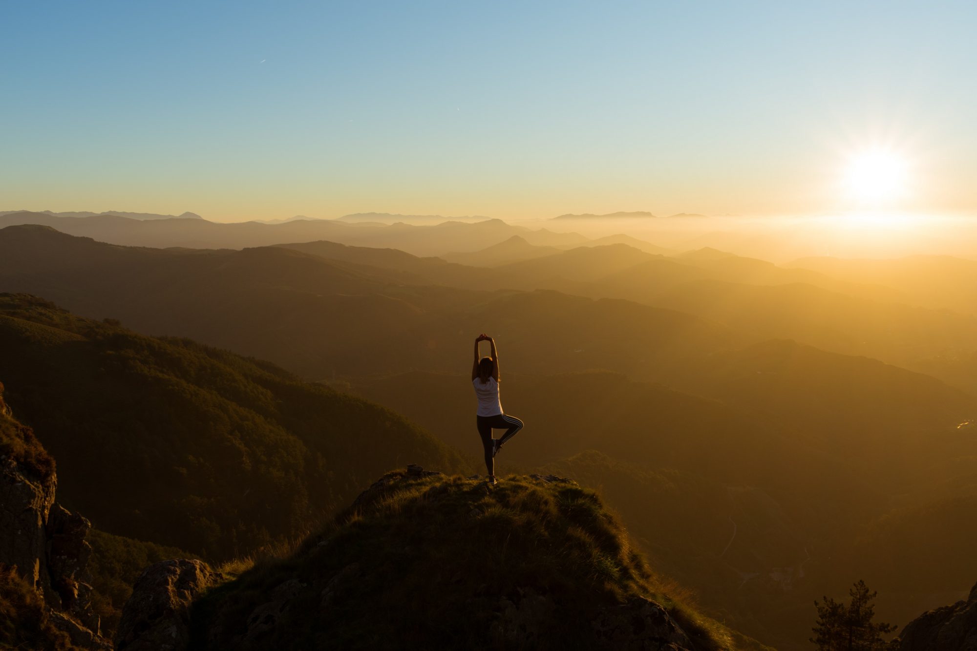 Une femme fait un figure de yoga sur un rocher tout en contemplant le coucher de soleil